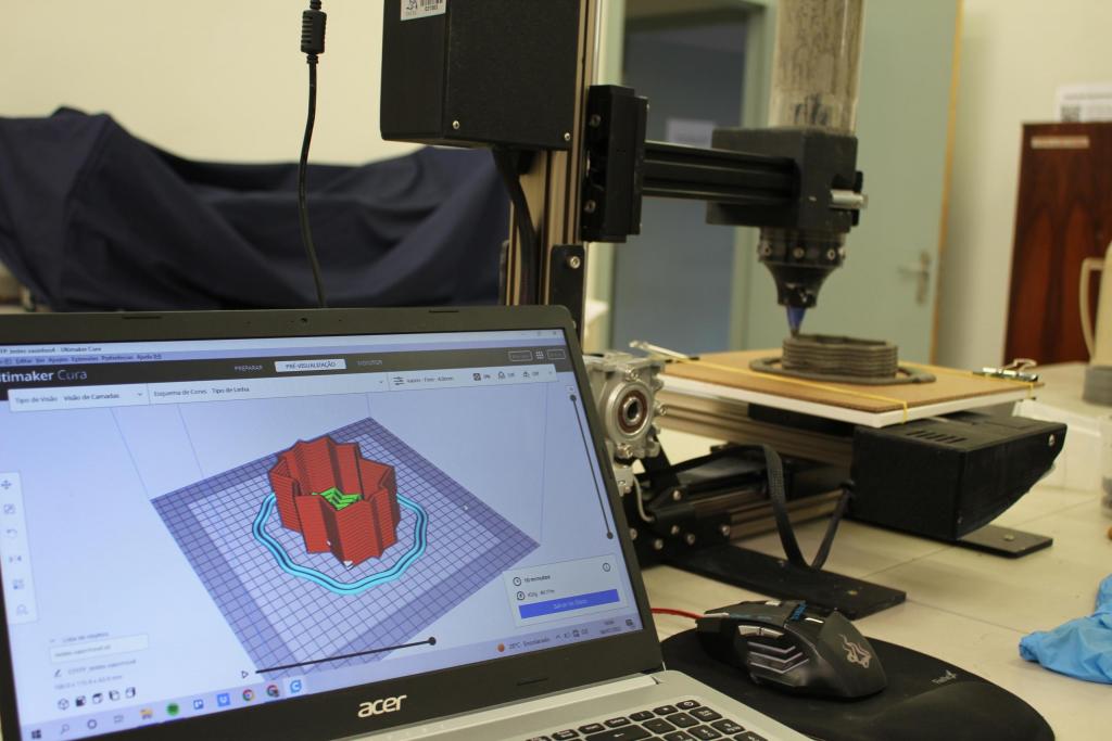 Fotografia horizontal colorida, em primeiro plano de um laboratório. Na extremidade direita da imagem,  um notebook preto,  aberto. Na tela, o programa que controla a impressora, com o modelo em 3D do molde de concreto nas cores vermelha, verde e azul. Em segundo plano está a impressora 3D. Ela é da cor preta, tem elementos metálicos e está sobre uma mesa branca. No fundo da imagem, elementos que compõem o laboratório. Na extremidade direita, um pano azul marinho que cobre alguns equipamentos. Ao lado, uma porta cinza, aberta com o corredor desfocado. Na parte esquerda da foto, um armário de madeira.
