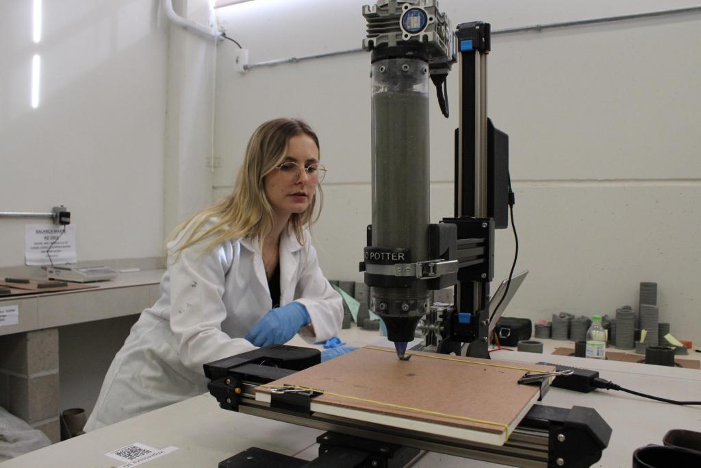 Fotografia horizontal, colorida, em plano médio, de uma mulher no laboratório. A mulher é branca, tem os cabelos loiros e soltos, veste um jaleco branco, usa luvas de plástico azul e óculos de grau na cor dourada. O olhar está direcionado para uma impressora 3D. A impressora 3D é preta, tem elementos metálicos e está sobre uma mesa branca. Na parte esquerda da imagem, uma mesa de azulejo branco, com uma balança e moldes de concreto em cima. À direita, moldes de concreto de tamanhos diferentes em cima de uma mesa branca. Ao fundo, estão as paredes brancas do laboratório.
