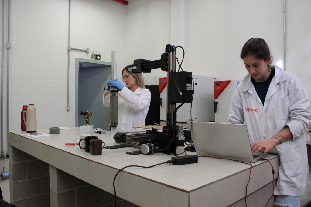 Fotografia horizontal, colorida em plano geral de duas mulheres em um laboratório. Na parte esquerda da imagem,  uma mulher branca, com cabelos loiros e soltos. Veste um jaleco branco e usa luvas de plástico azul e óculos de grau na cor dourada. Ela segura um tubo cinza. Ao lado dela, uma mulher branca, com cabelos castanhos presos em uma trança. Ela veste um jaleco branco com a logo do Grupos de Estudos em Materiais Sustentáveis na Construção (GEMASC) em laranja no peito. As mãos estão sobre um notebook cinza. O notebook está sobre uma mesa de azulejo branco. Na mesa, tem uma impressora 3D, na cor preta, um carregador de notebook, uma garrafa térmica, na cor bege, uma garrafinha de água, na cor laranja, e duas xícaras de café, nas cores preta e marrom. Ao fundo, as paredes brancas do laboratório e uma porta entreaberta, na cor cinza.