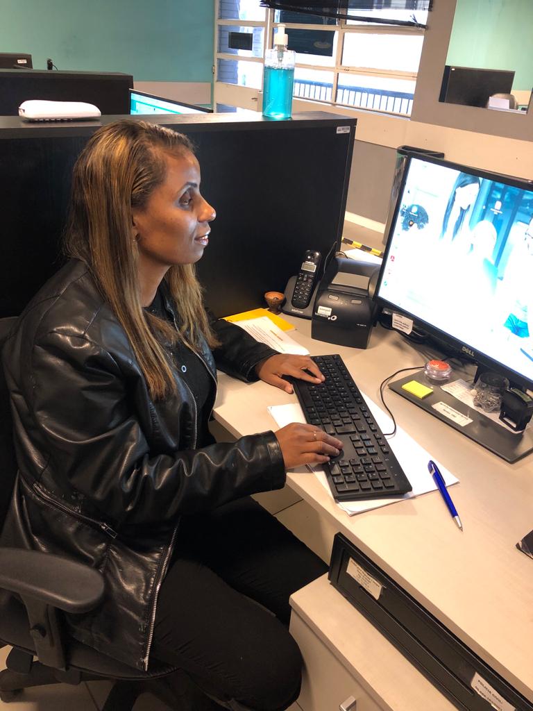  Fotografia vertical e colorida de Letícia, uma mulher negra sentada em frente a um computador. A fotografia está em plano geral fechado. Letícia está de perfil com uma expressão facial concentrada. Ela tem cabelos lisos, castanhos e com mechas loiras, na altura do peito. Veste roupas na cor preta: um casaco de couro sobre blusa com botão, e uma calça justa. O computador é preto e está com a tela ligada, mas não é possível definir o plano de fundo da tela do computador por conta da exposição alta. O computador está sobre a mesa de tampo bege claro. Também há um teclado, um telefone fixo e uma pequena impressora de notas, todos na cor preta, além de folha de papel e caneta azul. A cadeira em que ela está sentada é da cor preta. Atrás da mulher, divisórias pretas em formato horizontal. Ao fundo, retângulos de vidro, com nove divisórias  e acabamento em bege claro, além de paredes em tom de verde desbotado. 