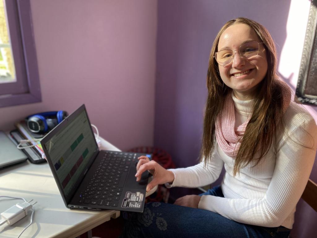 Fotografia quadrada, horizontal e colorida de uma mulher jovem sentada ao lado de um notebook, que sorri amplamente. Ela é uma mulher cis, branca, com rosto arredondado, olhos e boca finos, nariz médio e bochechas grandes. Tem cabelos lisos, loiros escuros, com comprimento na altura do peito e estão dispostos para a frente. O notebook é preto, está na posição diagonal e sobre a escrivaninha de cor clara. A tela tem informações, mas não é possível visualizar o conteúdo. Ela está com a mão direita sobre a parte do mouse do notebook. Ao fundo da imagem, a parede do quarto no ângulo em formato de “L”: a parede do lado esquerdo da imagem é roxo claro, e a do lado direito da imagem, na cor roxo escuro. Na extremidade superior esquerda da imagem, início de uma janela com aberturas na cor roxo escuro e com o vidro fechado Pela janela, entra a iluminação do dia..  À frente da janela, a escrivaninha com o notebook, um carregador de celular branco, fone de ouvido do tipo headset na cor preto e azul, e materiais de escritório. 