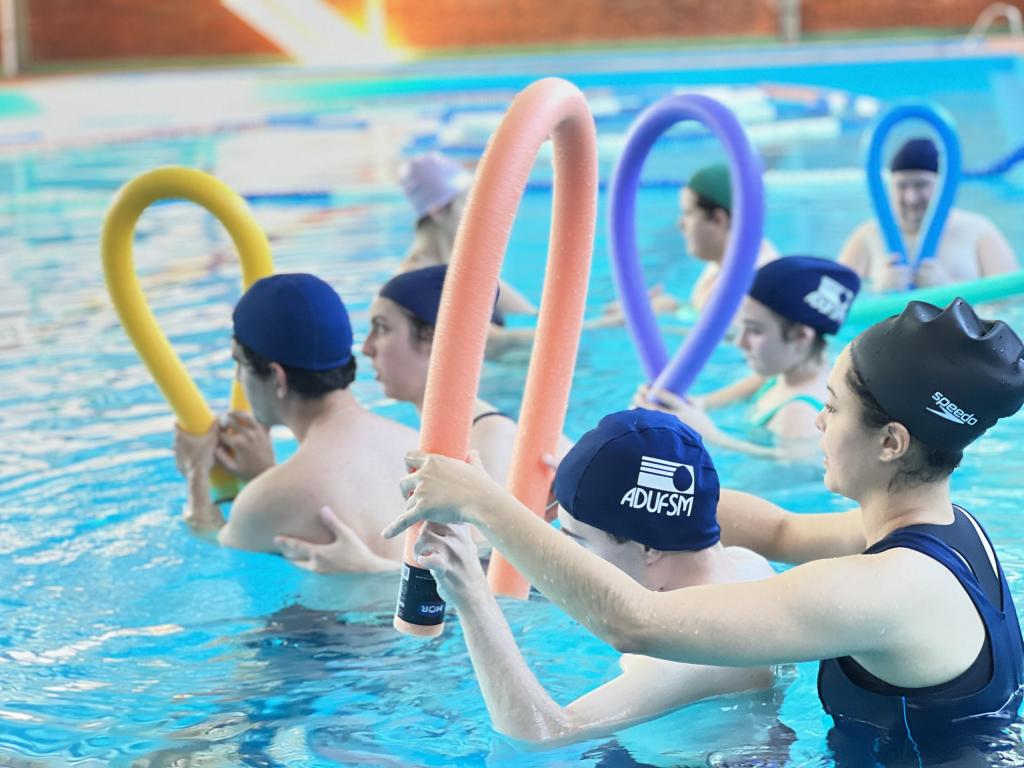 Fotografia horizontal e colorida de pessoas em uma piscina azul clara. São cerca de oito pessoas, que vestem roupas e toucas de natação em tons de azul marinho e seguram espaguetes de piscina coloridos, em tons de amarelo, azul e salmão. Em primeiro plano, duas pessoas em destaque, uma atrás da outra. A pessoa de trás segura o espaguete e auxilia o adolescente, que está na frente.Em segundo plano, cinco pessoas também vestem roupas de natação e seguram espaguetes de piscina; estão em dois grupos, um de dois e um de três pessoas. Ao fundo, em desfoque, a continuação da piscina, boias de piscina nas cores azul, roxa e amarelo, além da parede de tijolos a vista na cor marrom alaranjado.