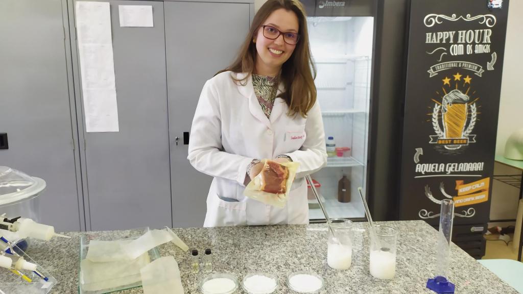 Fotografia horizontal e colorida de uma mulher de pé em um laboratório. Ela sorri, tem pele branca, rosto redondo, olhos escuros e cabelos lisos, compridos e na cor loiro escuro. Usa óculos de grau com armação e veste jaleco branco sobre blusa florida. Ela segura uma embalagem amarela e transparente, um pedaço de carne vermelha crua embalado a vácuo Ela está atrás de uma bancada de mármore cinza, na parte inferior da foto, em que há materiais de laboratório, como dois béqueres com um líquido branco, um tubo de ensaio e três placas de petri com pó branco dentro, além de outras embalagens abertas. Ao fundo, armários de cor cinza e uma geladeira de cor preta.