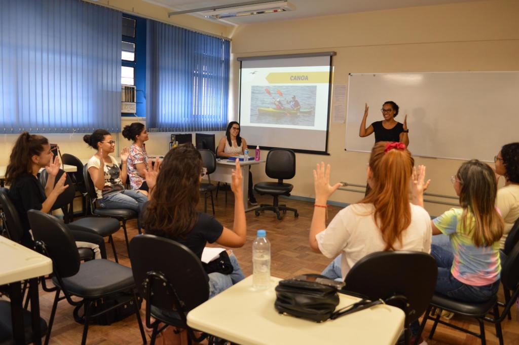 Fotografia horizontal e colorida de uma sala de aula com um grupo de pessoas sentadas em um semicírculo. Todas são mulheres. Em primeiro plano, as estudantes de costas estão sentadas em cadeiras pretas estofadas. Elas estão com as mãos levantadas e as palmas abertas, e praticam um sinal de Libras. A maioria tem pele branca e cabelos compridos em tons de castanho escuro, ruivo e loiro. Usam camisetas de manga curta nas cores preta, branca, creme, tie dye e florido. Em segundo plano, no centro do semicírculo, uma mulher em pé também está com as mãos levantadas. Ela sorri. Ela tem pele branca, tem cabelo preto, curto e liso; ela veste uma blusa preta e usa óculos. Atrás dela, quadro horizontal branco. Ao lado dela e na parte esquerda da imagem, uma mulher de pele branca sentada atrás de uma mesa bege com computador preto. Ela tem cabelo preto e liso, usa óculos e veste uma blusa branca. Atrás dela, tela de projeção de slides com uma imagem de canoa, sinal que o grupo está fazendo. Na extremidade esquerda da imagem, cortinas azuis. O fundo é uma parede branca.