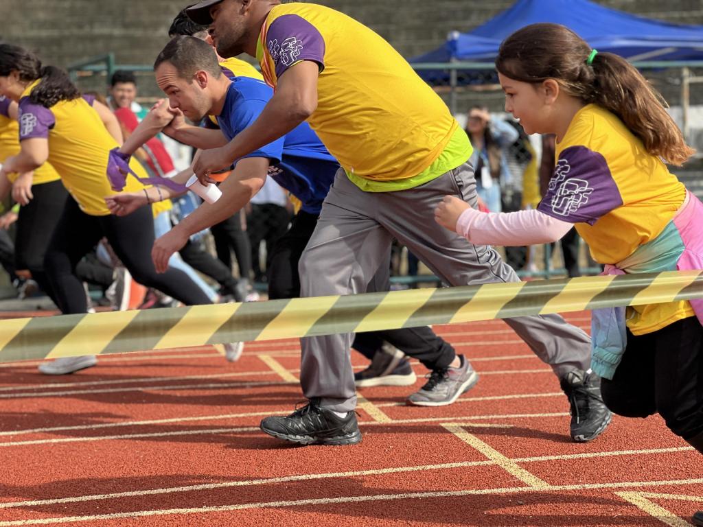 Foto na horizontal de uma corrida na pista de corrida da UFSM. Quatro pessoas se preparando para a largada da corrida em pista de corrida de cor vermelha. Menina em destaque, usa camiseta amarela com mangas roxas e blusa rosa por baixo, cabelo amarrado. Homem ao lado em posição de corrida também com blusa amarela com mangas roxas. Segundo homem da foto em posição de corrida com camiseta azul. 