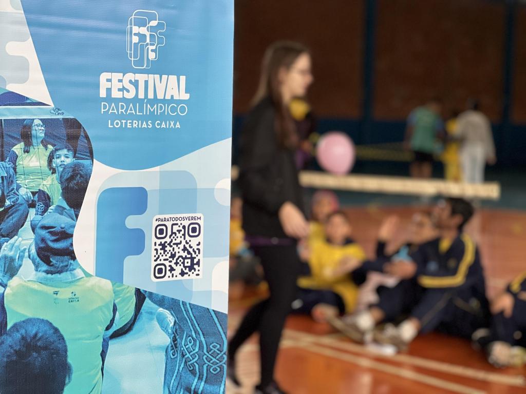 Foto em sentido horizontal. Banner em destaque frontal, nas cores azul, possui a escrita “Festival Paralímpico Loterias Caixa”, Qr code logo abaixo da escrita. Atrás do banner, posicionados em desfoque, cinco pessoas de frente para a rede de vôlei adaptado, com um balão inflável rosa, do outro lado da rede mulher de pé com blusa e calça preta.  