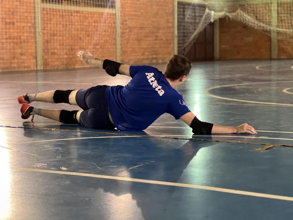 Foto na horizontal em quadra de futebol. Homem deitado de lado, com os braços estendidos em posição de defesa na prática do Goalball. Homem usa camiseta azul com a escrita “Atleta” nas costas, usa cotoveleira e tornozeleira pretas, tênis vermelho. Quadra de futebol em tijolos laranjas, chão azul com listras sinalizando a quadra em branco.