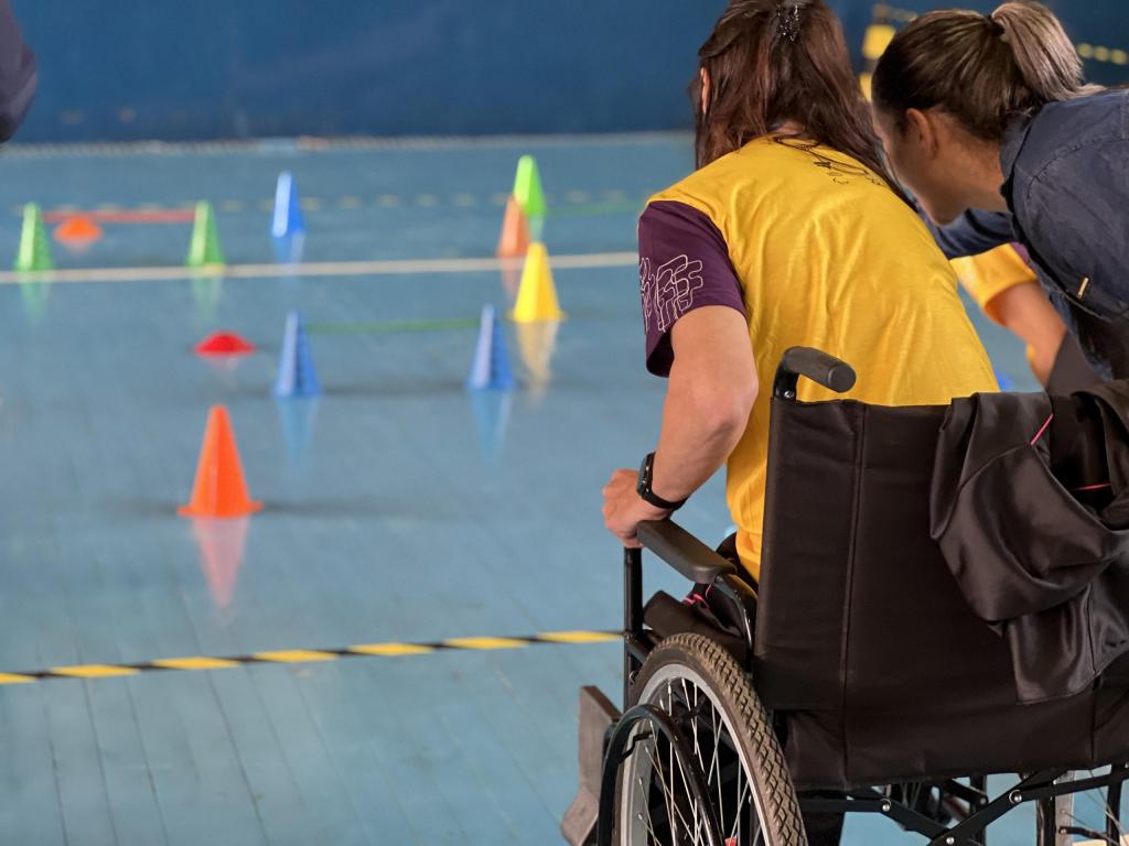 Foto na horizontal em quadra de jogos. Mulher em cadeira de rodas usa uma blusa amarela com mangas roxas. Joga bocha adaptada, em quadra azul claro com cones amarelos, azuis, verdes, vermelhos e laranjas. Posicionada ao seu lado, uma mulher com camisa azul escuro