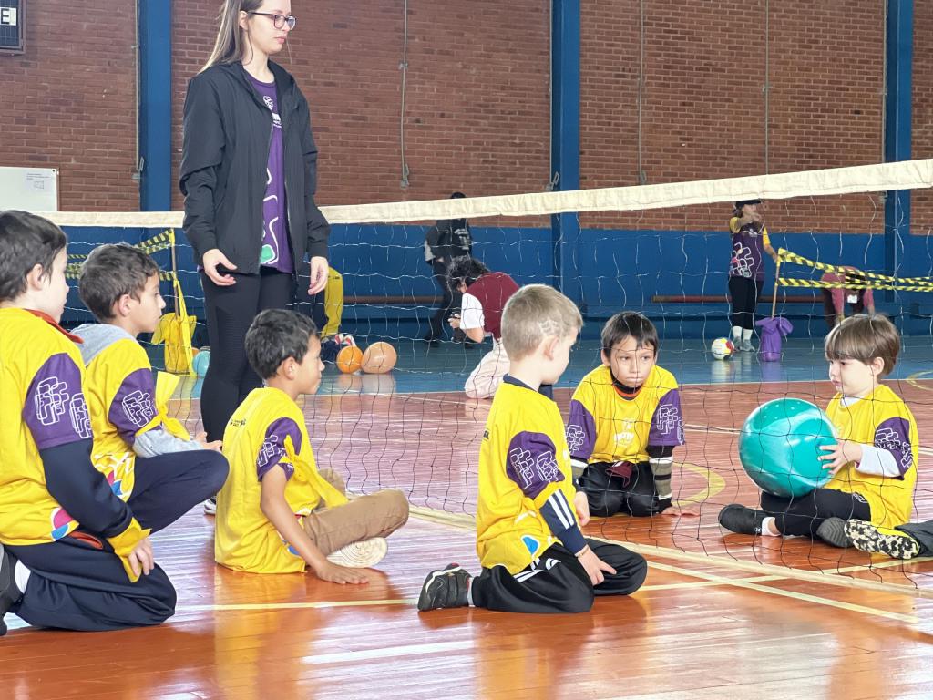 Foto em sentido horizontal representando a prática do vôlei sentado. Seis crianças sentadas no chão da quadra jogam o esporte. Quatro crianças do sexo masculino do lado esquerdo posicionadas de frente para rede, usam camiseta amarela com mangas roxas. Dois meninos do lado direito, posicionados do outro lado da rede, também com camisetas amarelas de manga roxa, um deles segura uma bola inflável verde. Quadra de futebol com chão laranja e azul, e sinalização em listras na cor branca. Rede de vôlei ao centro da quadra. Professora monitora em pé ao lado dos alunos, com camiseta roxa e casaco preto.