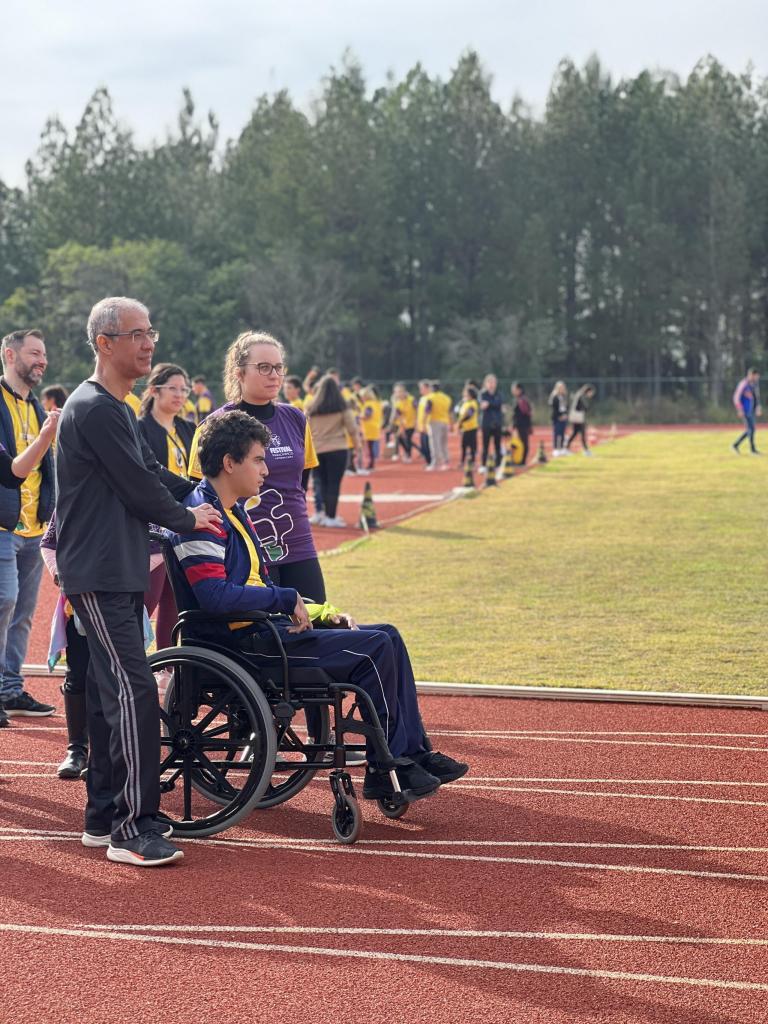 Foto na vertical na pista de corrida. Pista de corrida de cor vermelha com gramado de futebol ao centro. Homem de cadeira de rodas posicionado na pista pronto para a corrida, usa casaco azul com listras brancas e vermelhas. Homem de pé posicionado logo atrás, com a mão no ombro do homem sentado na cadeira de rodas, usa calça e camisa preta. Menina com camiseta roxa posicionada ao lado. Pessoas com camiseta amarela ao fundo em desfoque. 