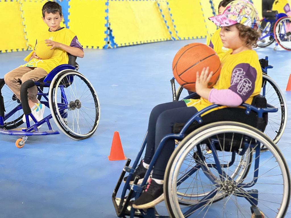 Foto na horizontal em quadra de basquete. Posicionados em quadra de basquete com chão azul e colchonetes amarelos ao lado. Menina em cadeira de rodas segurando uma bola de basquete laranja, com blusa amarela e mangas roxas, boné rosa e calça azul. Menino ao fundo em cadeira de rodas usa blusa amarela com mangas roxas, segura bola amarela.