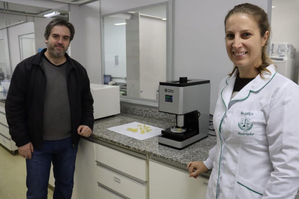 Fotografia horizontal colorida de um homem e uma mulher em um laboratório, em pé em frente a uma impressora 3D cinza e preta.  O homem está no lado esquerdo da fotografia. Ele tem pele branca, cabelo castanho, liso e curto, além de barba rala grisalha. Veste uma jaqueta preta sobre blusa de tricot em tom grafite e calça jeans escura. Ele está apoiado no tampo de granito cinza. A mulher está à direita do homem, tem pele branca, cabelo loiro escuro, liso, comprido e preso em um rabo de cavalo. Ela sorri e veste um jaleco branco sobre blusa preta. Também está escorada no tampo de granito. Atrás deles, em cima de uma bancada de granito, está a impressora 3D, cinza e retangular, pouco maior do que uma cafeteira e em formato parecido. Ao lado da impressora, vários chocolates no formato de animais em cima de uma folha de papel branco. Os chocolates são pequenos, tem formato de esfinge, tartaruga, lagarto e coelho e tem diferentes tons de branco e creme. Ao fundo, a parede é uma divisória cinza com uma janela.
