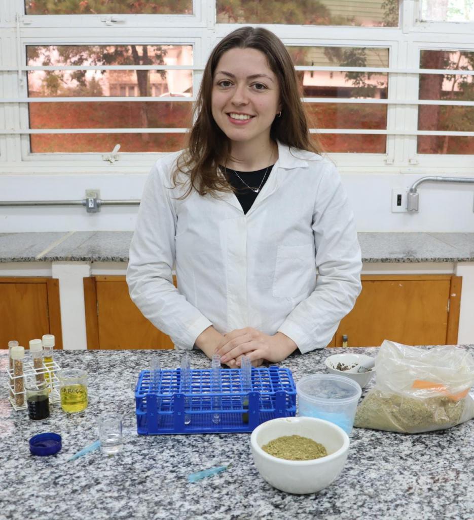 Fotografia vertical e colorida de uma mulher parada atrás de uma bancada de granito cinza em que há materiais de laboratório. Ela tem pele branca, cabelo castanho em tom médio, levemente ondulado e com comprimento na altura do ombro. Ela sorri amplamente, veste jaleco branco sobre camiseta preta e usa colar dourado com pingente. Está com as mãos apoiadas na bancada. Em sua frente,  amostras da oliva em óleo e em pó sobre uma bancada de granito dentro de tubos de ensaio, um pote e tigelas. Ao fundo, outra bancada com tampo de granito cinza, armários marrons, janelas com moldura branca e cano cinza, com entradas de tomada.
