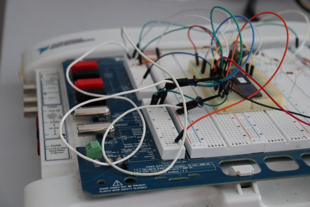 Fotografia horizontal e colorida de um protoboard com arduíno e fios nas cores vermelho, amarelo, azul, branco, verde e preto. O equipamento tem uma base fina e retangular. A camada de baixo é branca, a segunda, mais fina e em metal, é azul marinho, e a terceira é composta por blocos retangulares brancos com micro furos. Na terceira camada, os fios estão conectados. São vários fios, conectados em diferentes partes, e eles estão enrolados. Nas duas primeiras camadas, há informações textuais em letras pequenas, mas não são possíveis de serem lidas. A peça está sobre uma mesa branca.