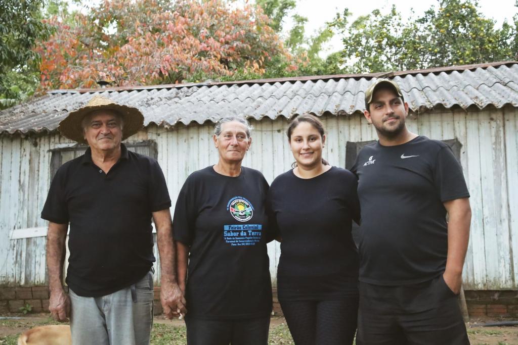 Fotografia horizontal e colorida  de quatro pessoas de pele branca, em pé em frente a uma casa velha de madeira. São duas mulheres no centro, e dois homens, um em cada extremidade. Eles sorriem levemente. No lado esquerdo está o Senhor Darci, de pele clara, estatura média e com aproximadamente 60 anos. Ele veste uma camisa gola polo preta, uma calça jeans escura e usa um chapéu de palha. Ele está de mãos dadas com Dona Maria, que está ao seu lado. Ela tem pele clara, estatura média e com aproximadamente 60 anos. Tem cabelos brancos presos em rabo de cavalo. Ela veste uma camiseta preta com uma estampa da logo do “Feirão Colonial” e uma leggin preta. Ao seu lado, está Ana Paula, mulher de pele clara, com cabelo castanho escuro preso em rabo de cavalo, estatura média e com aproximadamente 30 anos. Ela veste camiseta e leggin pretas. Por último, ao seu lado, está Jefferson, um homem de pele clara, estatura alta, e com aproximadamente 30 anos. Ele usa um boné verde escuro, veste camiseta e bermuda pretas. Jefferson está com a mão esquerda no bolso da bermuda. Atrás deles, a casa de madeira, que é de cor branca envelhecida e tem telhado de zinco. Ao fundo da imagem e acima da casa, está o topo de algumas árvores em tons de verde e laranja. O chão é de terra.