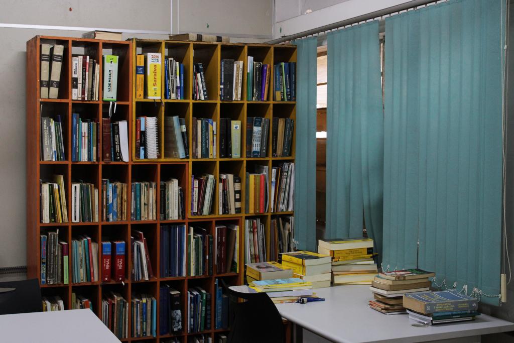 A fotografia horizontal e colorida retrata a biblioteca da CEU I. Uma sala com várias estantes de livros, mesas e cadeiras. Na parte superior, o teto apresenta pintura em listras largas nas cores azul, verde, amarelo, laranja e vermelho. Logo abaixo, a parede de fundo é lisa e clara, com três estantes de madeira repletas de livros e papéis empilhados, sendo que a estante da esquerda está mais desorganizada, com pilhas irregulares. No centro da sala há várias mesas retangulares brancas alinhadas, cercadas por cadeiras pretas de encosto vazadas. No canto direito, encostada na parede, há uma estante de nichos amarelos e marrons com livros, à frente de uma cortina vertical azul clara. Na parte inferior direita, outra estante verde aparece parcialmente, também carregada de livros. O piso é de madeira desgastada e há uma mesa com tampo de vidro próximo ao canto inferior esquerdo da imagem.