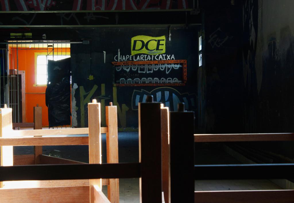 A fotografia é horizontal e colorida, ela retrata a antiga boate do DCE. O ambiente interno está escuro, com móveis de madeira empilhados, a única luz do cômodo vem das janelas de vidro. Na parte superior, a parede de fundo está coberta por tinta preta e grafites, com a sigla “DCE” em letras amarelas e pretas no centro, acompanhada das palavras “CHAPELARIA/CAIXA” em branco. Logo abaixo, há um balcão pequeno com moldura laranja e pichado. À esquerda, vê-se uma porta gradeada aberta, revelando uma sala pintada de laranja e com uma janela branca por onde entra luz. Na frente, ocupando a parte inferior, há mesas ou balcões de madeira virados de cabeça para baixo, recebendo feixes de luz. Ao fundo, a lateral direita permanece na penumbra.

