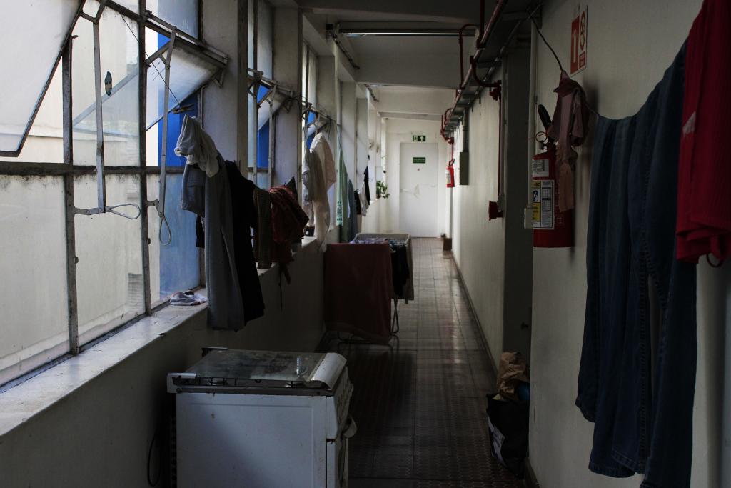 A foto na horizontal mostra um menino apoiado em uma janela, com o braço para fora, ele está observando a vista da cidade, onde ao fundo se encontram prédios e montanhas. Na imagem o estudante Robson, está na esquerda e se mostra de perfil encostado em uma janela, olhando para fora. Ele usa óculos e está vestindo um moletom cinza escuro com capuz branco, o cabelo é castanho escuro e está preso em um coque, Ainda ao fundo, vemos uma paisagem urbana da cidade de Santa Maria com prédios e, mais ao longe, montanhas cobertas por vegetação. A luz natural entra pela janela, iluminando levemente o rosto e a mão do homem. Se destacam tons de azul, verde, e creme na paisagem.
