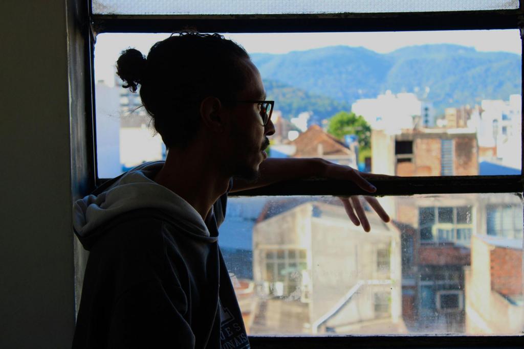 A foto na horizontal mostra um menino apoiado em uma janela, com o braço para fora, ele está observando a vista da cidade, onde ao fundo se encontram prédios e montanhas. Na imagem o estudante Robson, está na esquerda e se mostra de perfil encostado em uma janela, olhando para fora. Ele usa óculos e está vestindo um moletom cinza escuro com capuz branco, o cabelo é castanho escuro e está preso em um coque, Ainda ao fundo, vemos uma paisagem urbana da cidade de Santa Maria com prédios e, mais ao longe, montanhas cobertas por vegetação. A luz natural entra pela janela, iluminando levemente o rosto e a mão do homem. Se destacam tons de azul, verde, e creme na paisagem.
