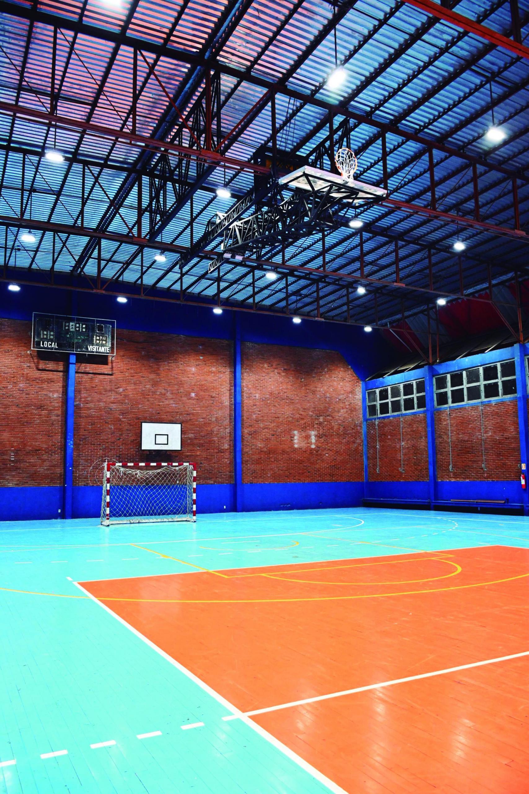 Fotografia vertical e colorida de um ginásio poliesportivo vazio à noite, iluminado por luzes artificiais. No chão, há linhas brancas que delimitam as áreas destinadas a diferentes modalidades esportivas, nas cores azul e laranja. Ao fundo, há uma goleira de estrutura de ferro nas cores vermelho e branco. Acima dela, há uma estrutura de cesta de basquete na cor branca com marcações pretas e, mais acima, a presença de um placar eletrônico. A estrutura de ferro do teto está exposta e há diversas lâmpadas de led fixadas nela, além de uma cesta de basquete. Ao fundo, paredes de tijolos à vista na cor marrom-claro com alguns detalhes em azul. Há janelas de vidro com esquadrias claras que mostram o lado de fora do ginásio, completamente escuro.
