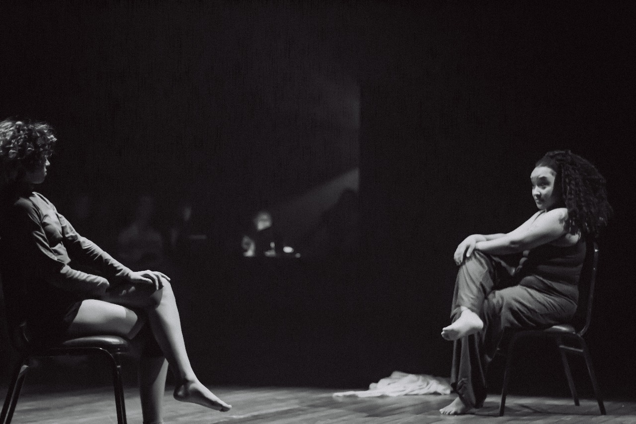 Fotografia retangular horizontal, preto e branco e em plano geral de duas mulheres sentadas em cadeiras no palco do Teatro Caixa Preta da UFSM. Elas estão com as pernas cruzadas, em diagonal uma em relação à outra. Estão na faixa etária dos 20 a 25 anos, têm pele negra, cabelos castanhos cacheados e olham uma para a outra. À esquerda, uma delas veste uma blusa de manga longa e um short. A outra, à direita, veste regata e calça. Ao fundo, há um tecido no chão e a mesa técnica do Teatro. O piso é feito de taco e as paredes são pretas.