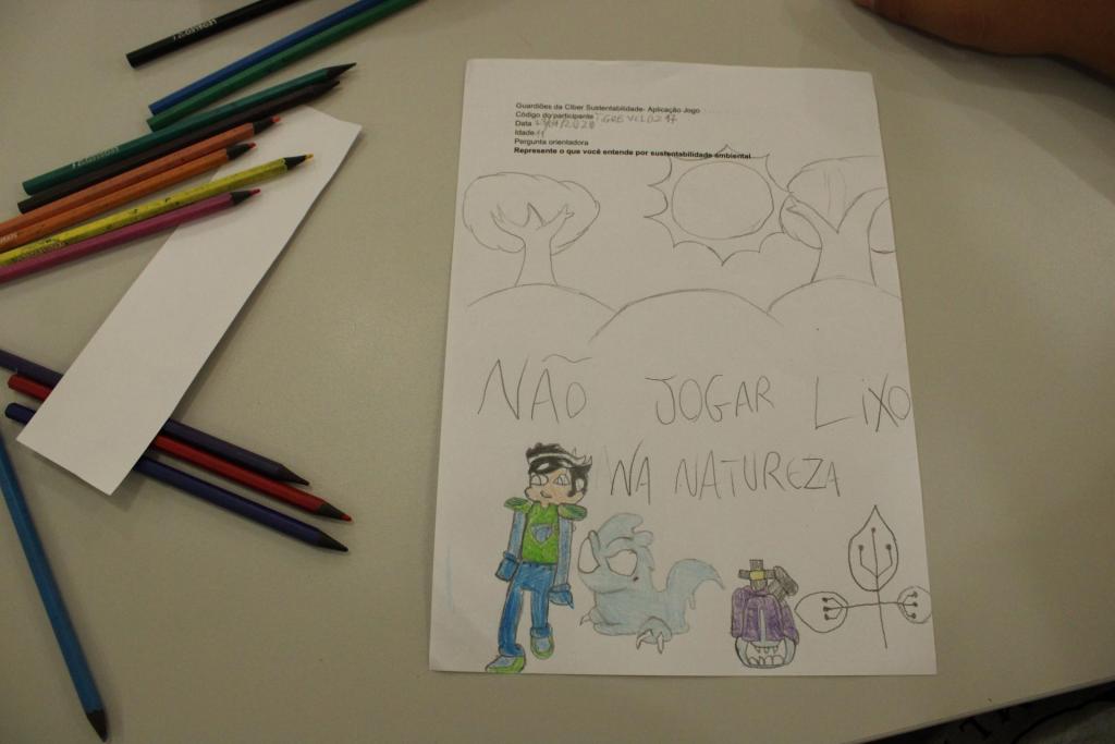 Foto horizontal e colorida de um desenho em uma folha sobre uma mesa cinza. Na folha estão desenhados um menino branco de olhos azuis, cabelo preto em formato de gota e armadura em verde e azul. Ao lado há um personagem bípede, similar a um anfíbio, de corpo azul e um robô de corpo roxo. Acima dos desenhos está escrito “Não jogar lixo na natureza”, seguido de duas árvores e um sol.. Do lado esquerdo do desenho, há uma régua branca e lápis de cor espalhados sobre a mesa ao redor da folha. O fundo da imagem é a mesa cinza.