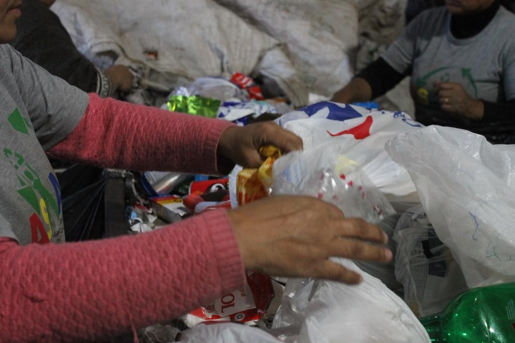 Fotografia horizontal e colorida. Em primeiro plano, duas pessoas aparecem parcialmente enquadradas enquanto separam resíduos recicláveis sobre uma esteira de triagem. A pessoa à esquerda veste camiseta cinza sobre blusa de mangas longas rosa e segura sacolas plásticas entre embalagens e materiais recicláveis. Ao fundo, outras pessoas trabalham na separação dos resíduos diante de grandes fardos de materiais prensados, em um galpão de reciclagem.