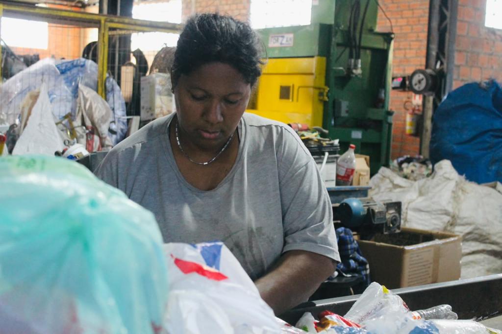 Fotografia horizontal e colorida. Em primeiro plano, uma mulher adulta, de pele negra e cabelos presos, veste camiseta cinza e realiza a separação de resíduos sobre uma esteira de triagem. Ela mantém o olhar voltado para os materiais à sua frente. Ao fundo, aparecem fardos de recicláveis, uma prensa hidráulica verde e amarela, caixas e equipamentos utilizados no processamento dos resíduos dentro do galpão.