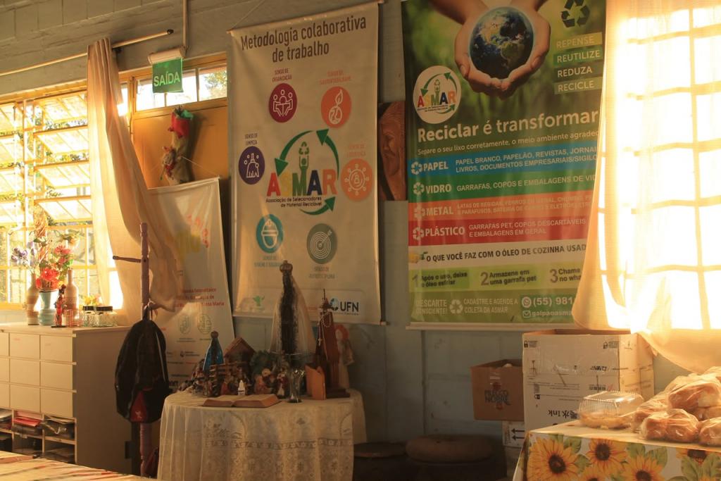 Fotografia horizontal e colorida. A imagem mostra o interior de uma associação de recicladores. Ao centro e à direita, dois grandes cartazes destacam orientações sobre sustentabilidade, reciclagem e separação correta de resíduos. À esquerda, há uma janela iluminada pela luz natural e uma mesa com toalha estampada, objetos decorativos e materiais diversos. No canto inferior direito, caixas de papelão e uma mesa com pães e embalagens completam o ambiente.