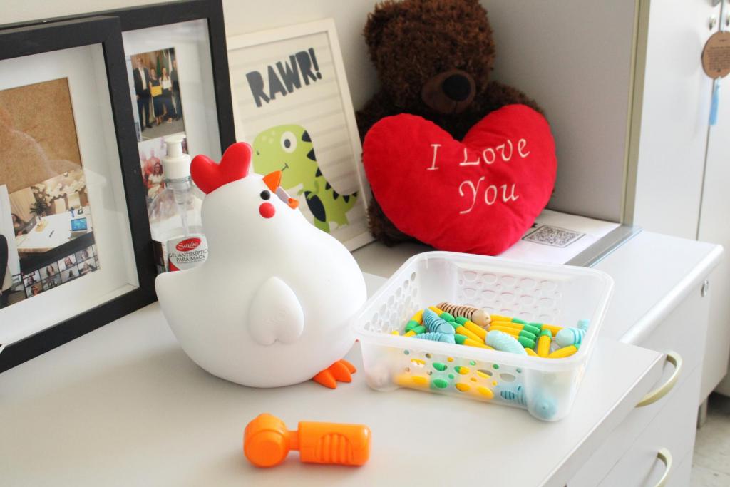 Foto horizontal e colorida de uma mesa com brinquedos e objetos decorativos, em um ambiente infantil ou de atendimento. No centro da imagem, há uma luminária branca em formato de galinha, com crista vermelha, bico laranja, bochecha vermelha e pés laranja. À direita da galinha, aparece uma cesta plástica transparente com peças coloridas em tons de amarelo, verde, azul e bege. Na parte inferior da imagem, sobre a mesa, há um brinquedo laranja em formato de martelo. À esquerda, aparecem porta-retratos com fotografias e um frasco de álcool em gel parcialmente escondido atrás da luminária. Ao lado do frasco de álcool em gel, ao fundo, há um quadro com a ilustração de um dinossauro verde e a palavra “RAWR!”. Ao lado aparece um urso de pelúcia marrom que segura uma almofada vermelha em formato de coração, com a frase “I Love You” escrita em branco. No fundo da imagem, há uma parede clara e um armário branco com puxadores.