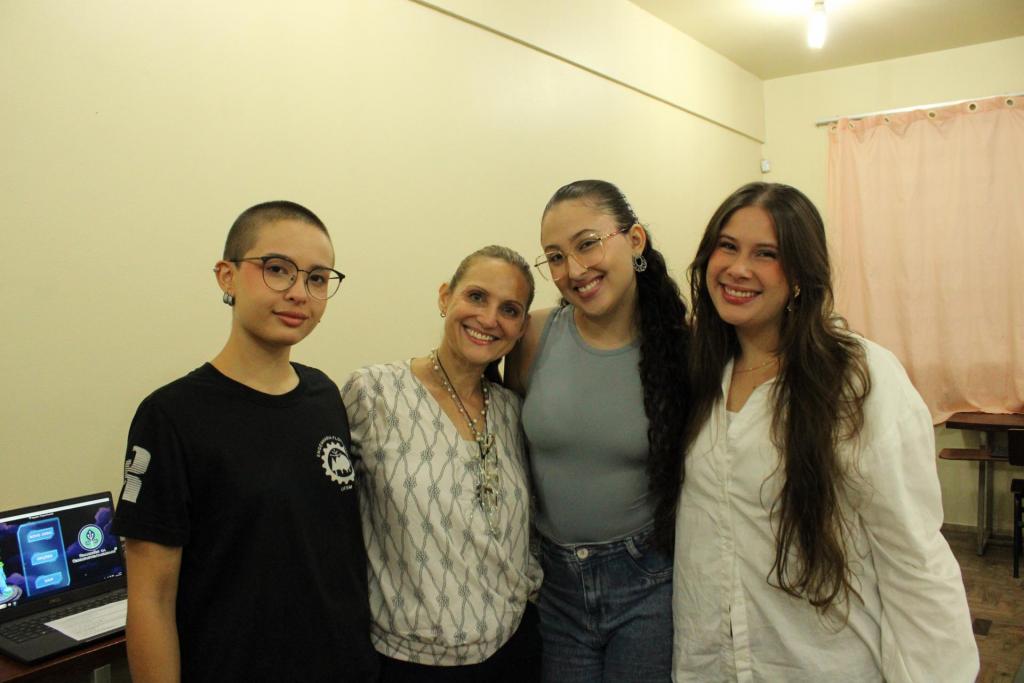 Fotografia horizontal e colorida de quatro mulheres em um laboratório de informática. À esquerda, Brenda de pele branca e de cabelo raspado. Ela usa óculos de armação preta e arredondada e veste camiseta preta. Ao lado, Eliane de pele branca de estatura baixa, cabelo loiro preso para trás. Ela usa um colar de pérolas e veste blusa branca. Em seguida está Luiza, uma mulher jovem de pele branca, estatura alta, cabelo cacheado, comprido e preto com a metade presa. Ela óculos de armação clara e gateada e veste regata cinza com jeans azuis. À direita está Rafaela, uma mulher jovem de pele branca, que tem cabelos castanho claros, compridos e lisos. Ela veste camisa social branca. Ao fundo, alguns notebooks sobre mesas marrons e a parede bege.