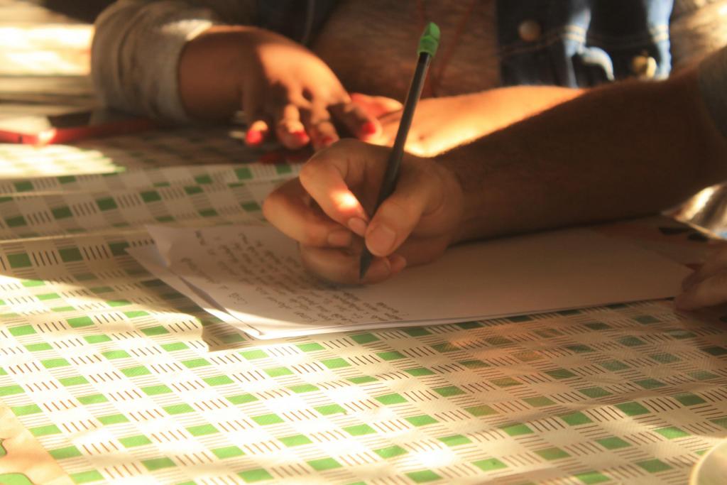 Fotografia horizontal e colorida de um estudante que escreve em uma folha de papel enquanto recebe acompanhamento da bolsista do projeto, durante uma atividade pedagógica. A fotografia tem enquadramento fechado e destaca as mãos do estudante e da bolsista. O foco está na mão do aluno, de pele parda, que usa camiseta cinza e segura um lápis preto com borracha verde sobre um papel branco com texto manuscrito. Atrás dele estão as mãos de outra pessoa, de pele negra e com as unhas pintadas de rosa. Ela veste jaqueta jeans com as mangas cinzas. Ambos apoiam as mãos sobre uma toalha de mesa estampada com quadrilhos verdes. A iluminação natural destaca a cena e produz sombras sobre a toalha a partir das mãos..