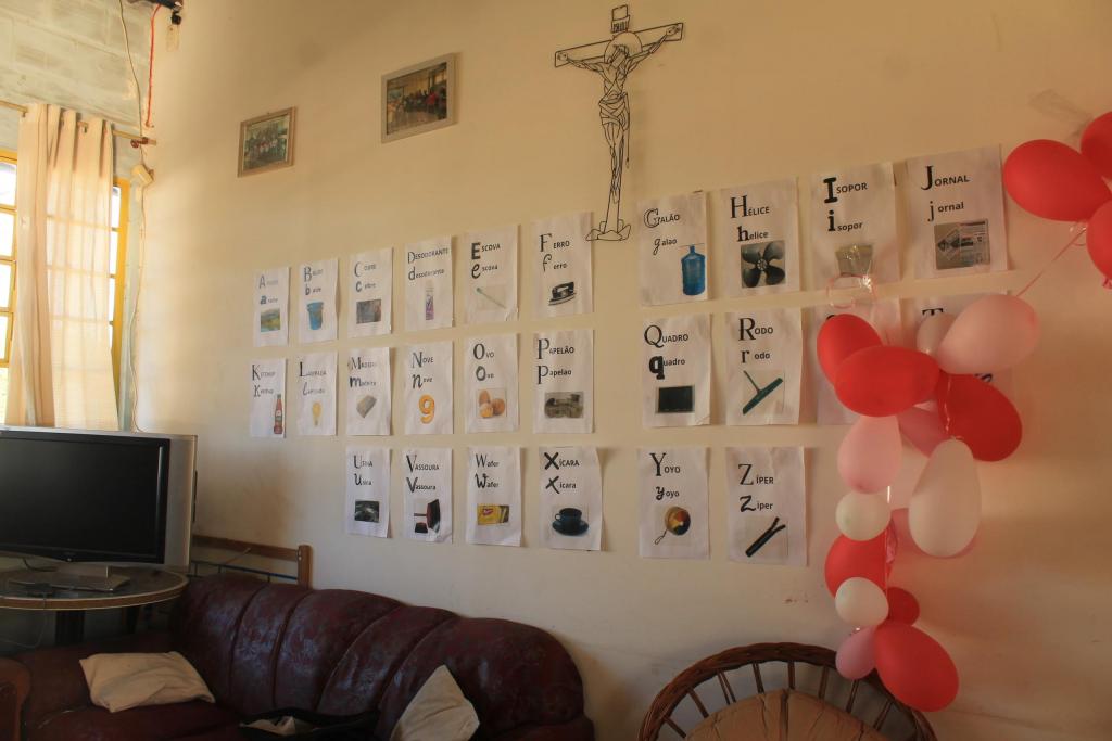 Fotografia horizontal e colorida de uma parede branca com cartazes e imagens que correspondem às letras do alfabeto. Organizados na sequência alfabética, os materiais apresentam palavras e figuras associadas a cada letra, como o x que é associado à palavra xícara. Do lado direto da imagem e dos cartazes, estão cerca de 18 balões nas cores vermelho, branco e rosa claro. Acima dos cartazes, há uma imagem representativa de Jesus Cristo e, ao lado esquerdo, dois quadros com imagens indistintas. Abaixo dos cartazes, aparecem cortinas de uma janela e móveis uma televisão,um sofá e uma poltrona. As paredes são bege.