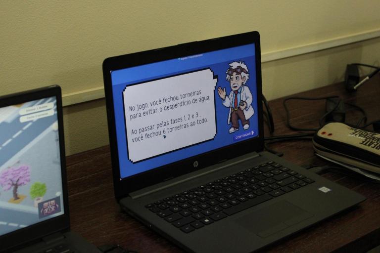 Foto horizontal e colorida de um notebook, sobre uma mesa de madeira, que mostra a tela de um jogo com ilustrações em pixel art. A tela do notebook é azul com o desenho de Na tela do notebook, um homem idoso, de bigode e cabelos brancos, que veste um jaleco, camisa social azul, gravata vermelha e um par de óculos de aviador. Ele dá instruções a partir de um balão de texto branco posicionado à sua esquerda. No canto inferior esquerdo, outro aparelho similar exibe o mesmo jogo. No canto superior direito, cabos e fios se espalham na mesa ao redor de um estojo branco. Ao fundo, a parede é bege.