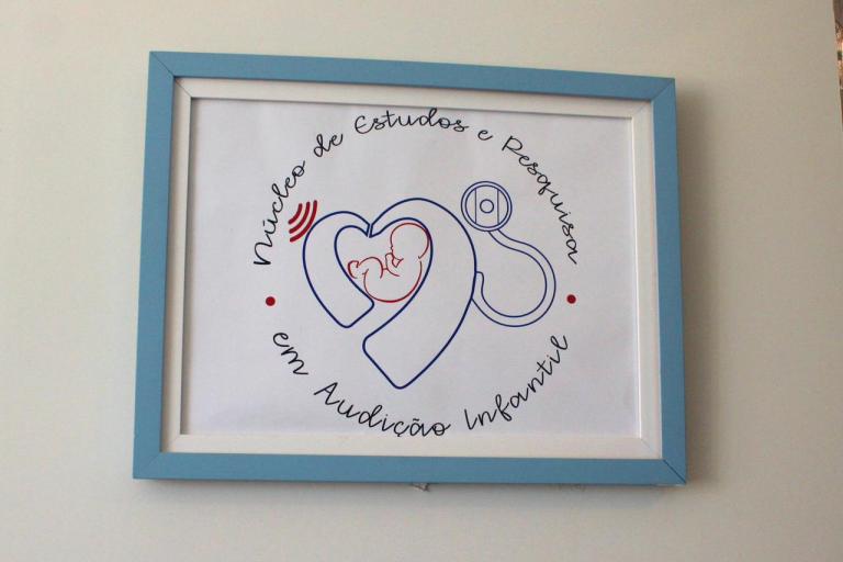 Foto horizontal e colorida de um quadro com a identificação do Núcleo de Estudos e Pesquisas em Audição Infantil. No centro da imagem está o quadro retangular, que tem moldura azul clara e fundo branco. Dentro dele, aparece um desenho formado por linhas azuis e vermelhas: à esquerda, há um coração com a ilustração de um bebê no interior; à direita, o desenho se conecta a um estetoscópio. Ao redor da ilustração, está escrito “Núcleo de Estudos e Pesquisas em Audição Infantil” em formato circular e letra cursiva. Também aparecem pequenos pontos vermelhos e linhas vermelhas que lembram ondas sonoras. Ao fundo, há uma parede clara.