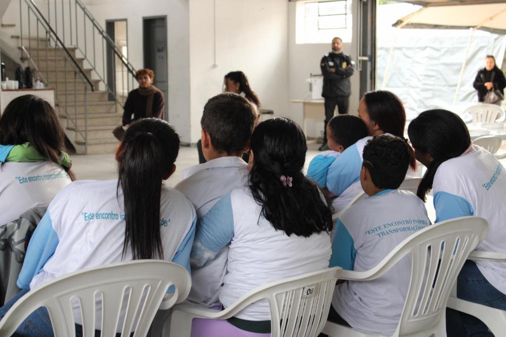 Fotografia horizontal e colorida de participantes do Projeto Inspira que assistem a uma apresentação durante encontro do grupo. Em primeiro plano, de costas, cinco mulheres e três crianças estão sentadas em cadeiras plásticas brancas, voltadas para a apresentação. Duas crianças estão sentadas no colo de mulheres e uma ocupa uma cadeira plástica. Todos vestem camisetas brancas com mangas azul-claras e a frase “Este encontro inspira transformações” estampada nas costas. Ao fundo, o ambiente amplo apresenta paredes brancas, escada à esquerda e abertura à direita, por onde entra iluminação natural. Cerca de quatro pessoas estão ao fundo, sendo duas sentadas no centro e duas de pé no lado direito. Uma das pessoas de pé é um homem que veste farda policial e está próximo à porta.