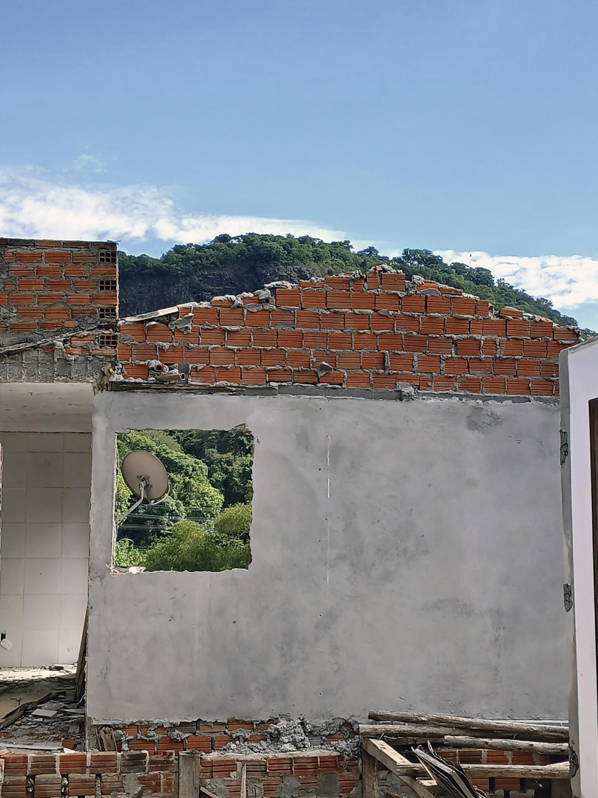 Fotografia vertical colorida de uma casa parcialmente destruída após danos estruturais. Ao centro da imagem, uma parede cinza com revestimento de cimento permanece de pé. Acima da parede, estrutura de tijolos alaranjados aparentes forma um triângulo que seria parte do telhado. Na parede cinza, há uma abertura quadrada onde antes ficava uma janela. Abaixo, a estrutura continua com tijolos alaranjados aparentes. Em primeiro plano, pedaços de madeira e restos de materiais de construção estão espalhados e empilhados junto à base da parede. Pela abertura da antiga janela, aparece uma antena parabólica instalada na área externa. Ao fundo, vegetação densa e verde ocupa a paisagem. Acima, céu azul-claro com poucas nuvens brancas.