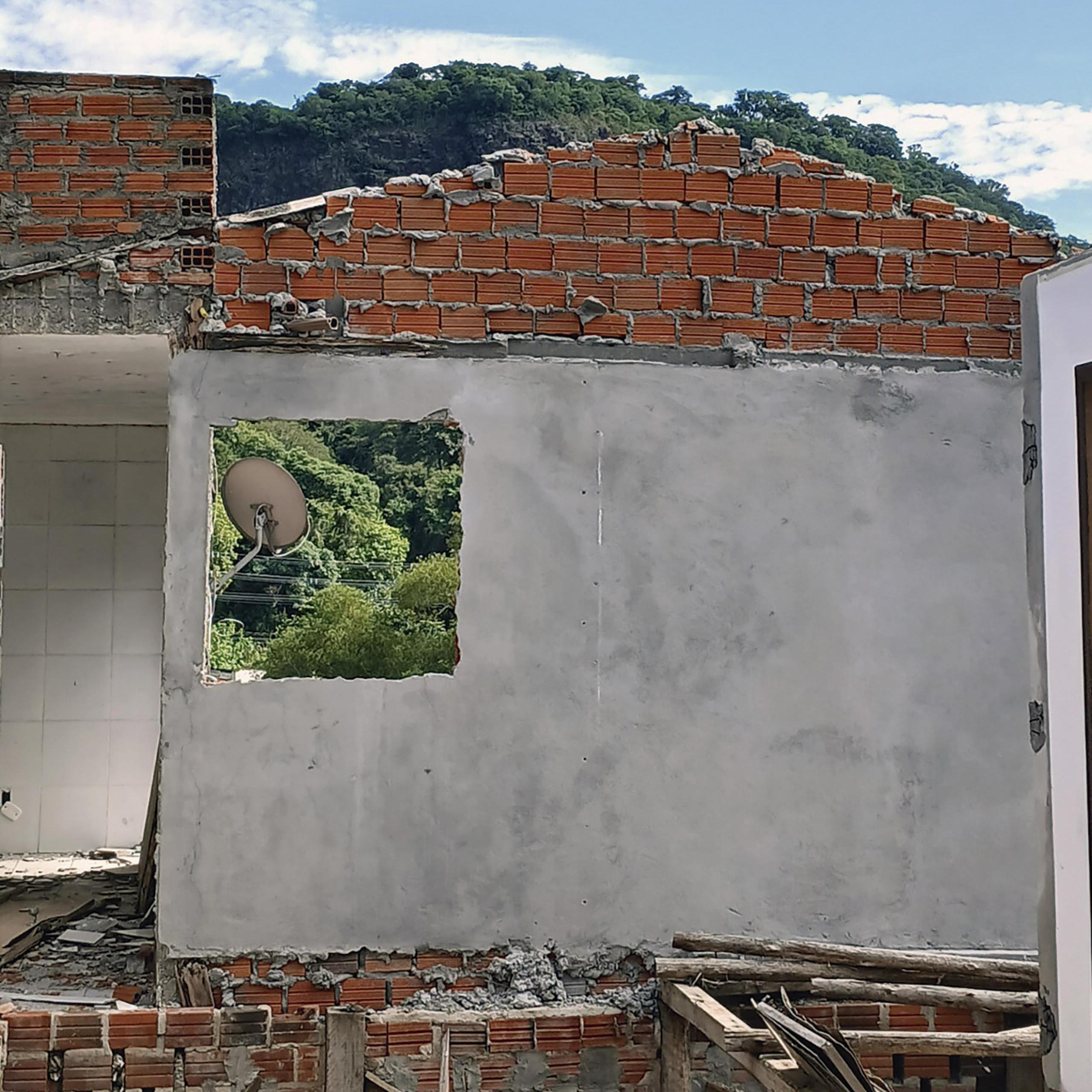 Fotografia vertical colorida de uma casa parcialmente destruída após danos estruturais. Ao centro da imagem, uma parede cinza com revestimento de cimento permanece de pé. Acima da parede, estrutura de tijolos alaranjados aparentes forma um triângulo que seria parte do telhado. Na parede cinza, há uma abertura quadrada onde antes ficava uma janela. Abaixo, a estrutura continua com tijolos alaranjados aparentes. Em primeiro plano, pedaços de madeira e restos de materiais de construção estão espalhados e empilhados junto à base da parede. Pela abertura da antiga janela, aparece uma antena parabólica instalada na área externa. Ao fundo, vegetação densa e verde ocupa a paisagem. Acima, céu azul-claro com poucas nuvens brancas.