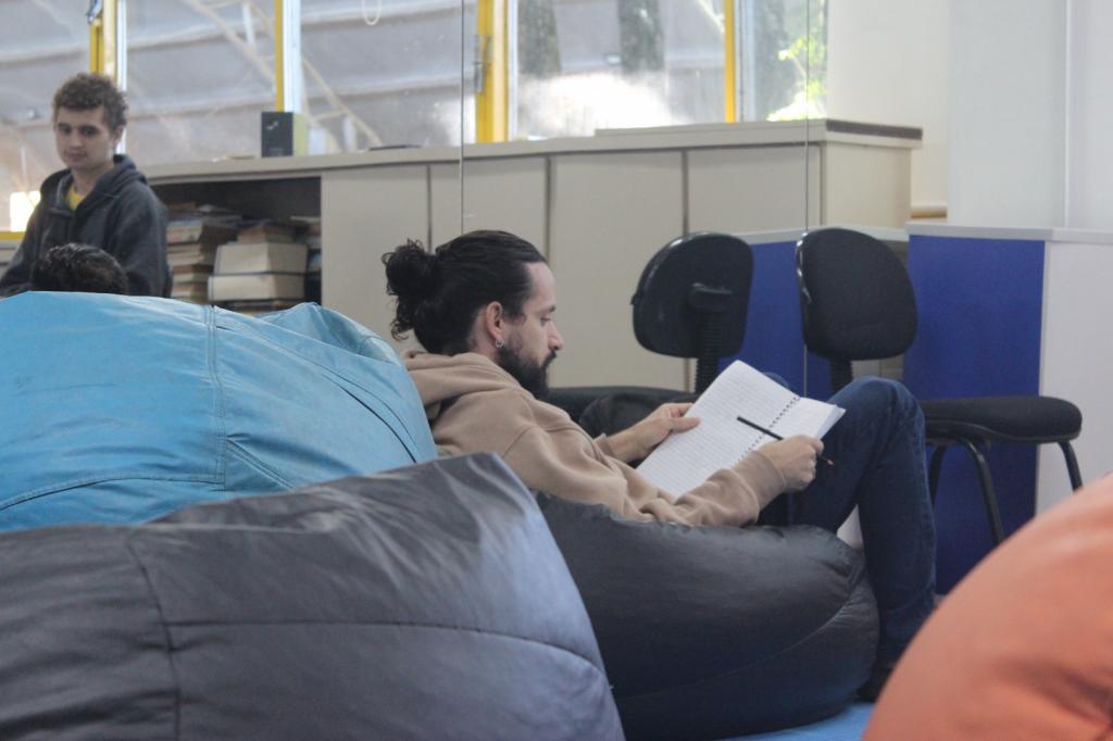 Foto horizontal e colorida de Davi sentado em um pufe preto com um livro nas mãos. Ele é um homem de pele branca, cabelos castanho-escuros compridos, presos em um coque, e usa barba da mesma cor. Ele veste moletom bege e calça jeans escuro. Em volta dele, há outros pufes grandes nas cores cinza-escuro, azul claro e laranja, que preenchem a parte inferior e esquerda da cena.  Atrás dele há duas cadeiras de escritório pretas, um balcão baixo bege com livros empilhados e grandes janelas de vidro ao fundo. À esquerda e em desfoque está outro rapaz, de pele branca, cabelos castanho-claros e cacheados e moletom cinza escuro. Ele observa a cena.
