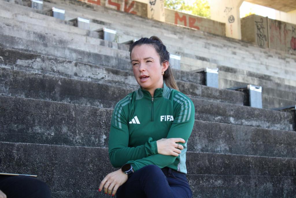 Foto horizontal e colorida de Maíra sentada em uma arquibancada. Posicionada no centro da imagem, Maíra é uma mulher de pele branca, cabelos castanhos com mechas loiras presos em um rabo de cavalo. Ela usa brincos de argola pequenos e dourados e tem as unhas vermelhas. Usa casaco verde do tipo esportivo, com listras brancas e zíper na gola, com a palavra FIFA e símbolo da marca Adidas no peito, e calça jeans azul marinho. Usa relógio preto no pulso esquerdo. As pernas e os braços estão cruzados em frente ao corpo. A expressão facial é séria e o olhar se direciona para a lateral. Está com a boca ligeiramente aberta e os olhos semicerrados. Ao fundo, na parte superior da imagem, aparecem estruturas de concreto de uma arquibancada, com algumas pichações nas paredes e áreas iluminadas pela luz natural. Atrás de Maíra há degraus de concreto cinza que ocupam grande parte do cenário.