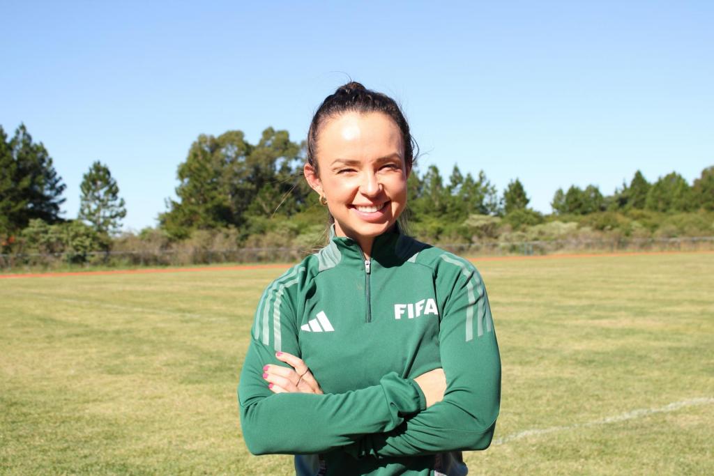 Foto horizontal e colorida de Maíra sorridente em um campo de futebol. Posicionada no centro da imagem, ela é uma mulher de pele branca, cabelos castanhos com mechas loiras presos em um rabo de cavalo. Usa brincos de argola pequenos e dourados e tem as unhas vermelhas. Ela usa um casaco verde do tipo esportivo, com zíper na gola e listras brancas, com a palavra FIFA e um logotipo da Adidas no peito, além de calça jeans azul marinho. Maíra está com os braços cruzados em frente ao corpo. No fundo, há um campo de futebol com gramado verde e linha branca de campo, goleira branca, árvores e céu azul.