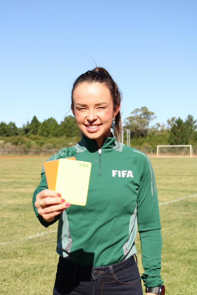 Foto vertical e colorida de Maíra em um campo de futebol. Posicionada no centro da imagem, Maíra está com expressão sorridente. Ela é uma mulher de pele branca, cabelos castanhos com mechas loiras, presos em um rabo de cavalo. Ela usa brincos de argola pequenos e dourados e tem as unhas vermelhas. Usa casaco verde do tipo esportivo, com zíper na gola e listras brancas, com a palavra FIFA no peito , e calça jeans azul marinho. Maíra está com o braço estendido à frente e segura um par de cartões nas cores amarelo e vermelho fluorescentes. No fundo, um campo de futebol com gramado verde e linha branca de campo, goleira branca, árvores e céu azul.
