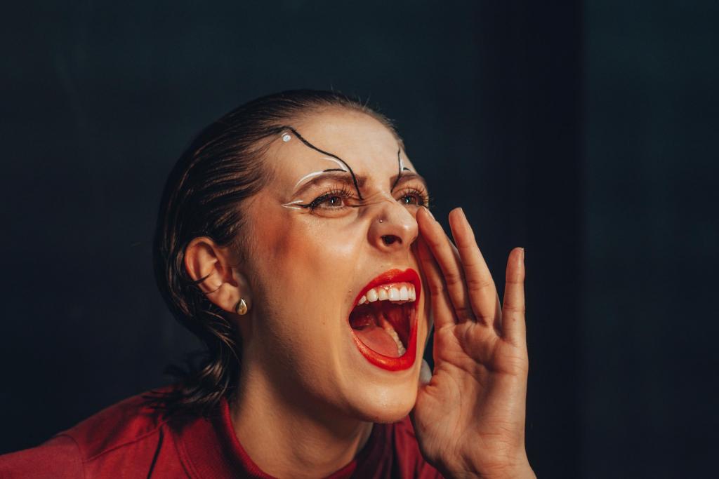 Fotografia retangular horizontal colorida de Sofia Lopes Dotto em cena artística. Ao centro da imagem, ela aparece enquadrada dos ombros para cima. Sofia está com o rosto voltado para a direita, boca aberta e mão direita posicionada ao lado da boca. A expressão é intensa e enérgica. Ela usa maquiagem elaborada, com batom vermelho, delineador claro ao redor dos olhos e elementos decorativos que se estendem pela testa. Os cabelos estão penteados para trás, com aparência úmida. Ela veste roupa vermelha em tom semelhante ao da maquiagem. Ao fundo, uma área escura e desfocada ocupa a imagem. Fotografia retangular horizontal colorida de Sofia Lopes Dotto em cena artística. Ao centro da imagem, ela aparece enquadrada dos ombros para cima. Sofia está com o rosto voltado para a direita, boca aberta e mão direita posicionada ao lado da boca. A expressão é intensa e enérgica. Ela usa maquiagem elaborada, com batom vermelho, delineador claro ao redor dos olhos e elementos decorativos que se estendem pela testa. Os cabelos estão penteados para trás, com aparência úmida. Ela veste roupa vermelha em tom semelhante ao da maquiagem. Ao fundo, uma área escura e desfocada ocupa a imagem. 