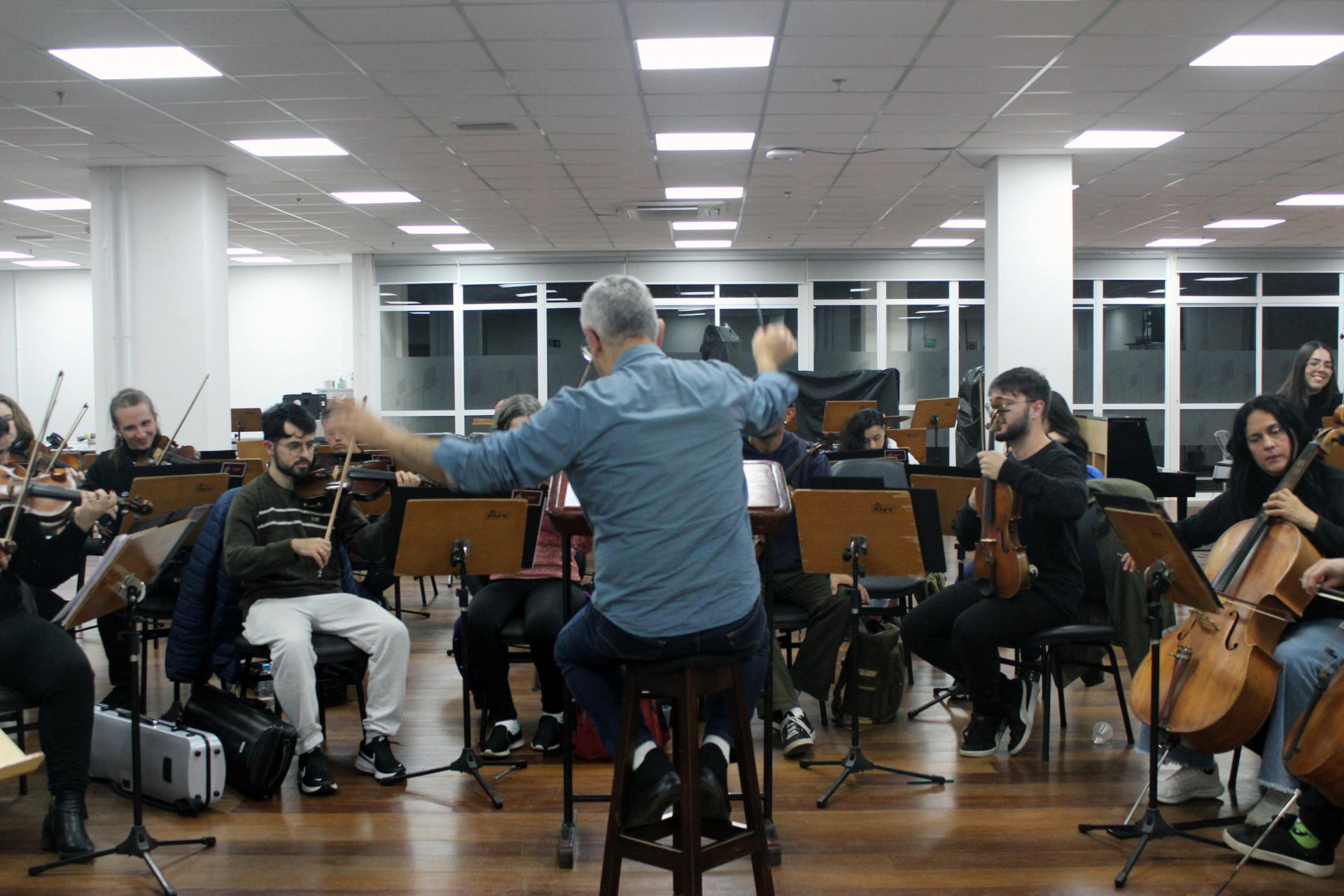 Fotografia horizontal colorida de ensaio de orquestra em sala de ensaio, com o maestro Cláudio Antonio Esteves e músicos em apresentação. Em primeiro plano, ao centro, está o maestro de pele branca sentado de costas em um banco alto de madeira escura. Ele tem cabelos grisalhos curtos e veste camisa azul-clara de mangas longas dobradas até os antebraços e calça escura. Com os braços erguidos, conduz a orquestra à frente. Ao fundo, doze músicos ocupam o restante da imagem e executam peças com instrumentos de corda e outros instrumentos orquestrais. Eles estão sentados atrás de estantes de partituras de madeira sobre tripés pretos.