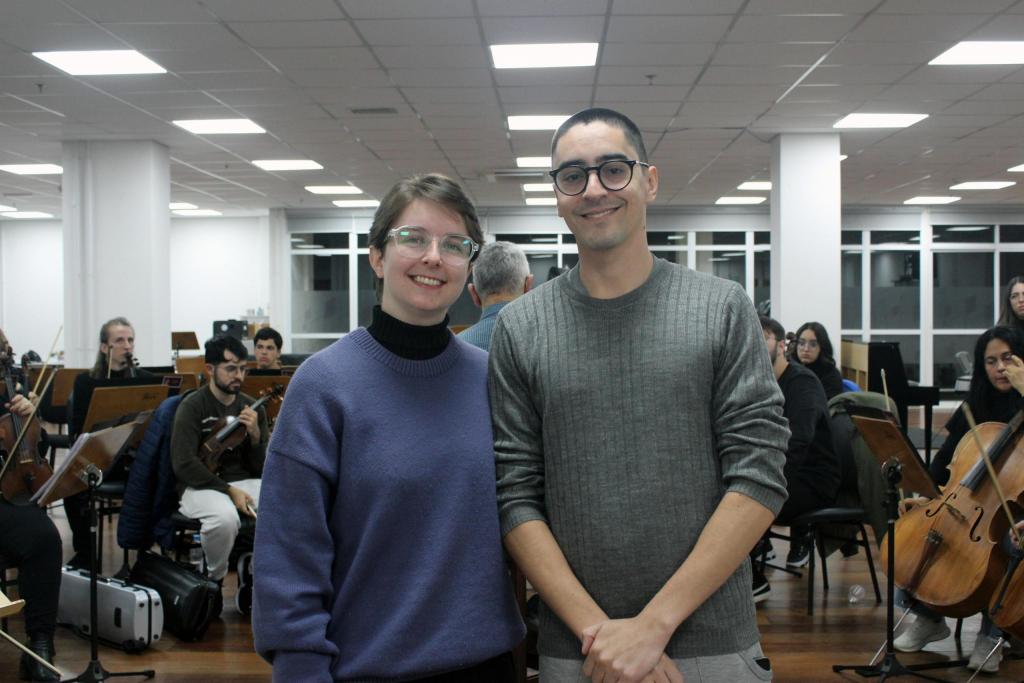 Fotografia horizontal colorida da flautista Gabriela Sara Griebeler e do clarinetista Eder Roberto de Oliveira lado a lado. Ao centro da imagem, ambos estão em pé e enquadrados da cintura para cima. A mulher tem pele branca, cabelos curtos e claros, usa óculos com armação transparente e veste blusão azul. Ao lado dela, um homem de pele branca tem cabelo raspado, usa óculos com armação preta e veste blusão cinza com mangas puxadas. Ele apoia a mão esquerda sobre a direita à frente do corpo. Ao fundo, os músicos permanecem sentados com instrumentos musicais nas mãos. Eles têm expressões concentradas e alguns olham para as estantes de partituras na frente deles. Atrás dos músicos, há uma janela ampla de vidros em meio a paredes e vigas brancas.