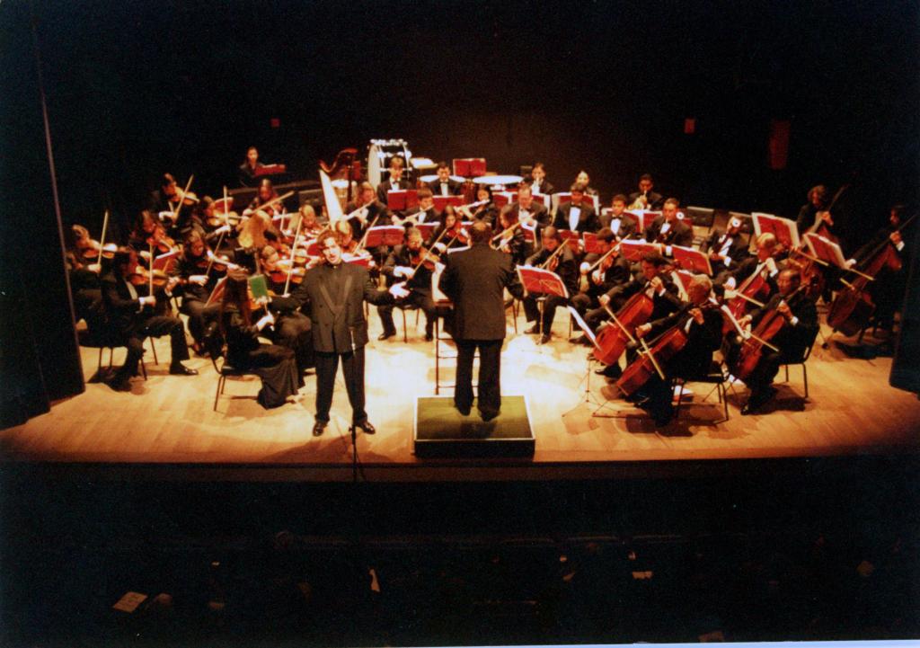 Fotografia horizontal colorida de apresentação da orquestra sinfônica em um palco iluminado de madeira clara. Em primeiro plano, ao centro, um solista está de frente para o público com os braços abertos. Ao lado dele, o maestro Enio Guerra está de costas sobre uma plataforma de regência com bordas brancas. Atrás do solista e do maestro, os musicistas, em sua maioria homens, estão sentados em frente a estantes de partituras em um semicírculo. Todos seguram instrumentos de cordas e vestem ternos e calças pretas. À esquerda da imagem, musicistas sentadas executam violinos e violas. À direita, integrantes da seção de violoncelos e contrabaixos permanecem sentados com os instrumentos posicionados à frente do corpo. Ao fundo, músicos de sopro e percussão ocupam fileiras organizadas atrás de estantes de partituras vermelhas. O palco tem piso de madeira clara e iluminação direcionada aos integrantes da orquestra.
