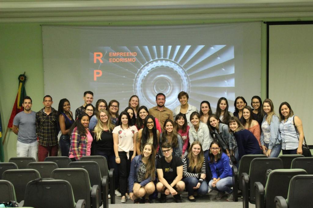 Diversas pessoas em pé em frete ao palco e a uma tela projeção