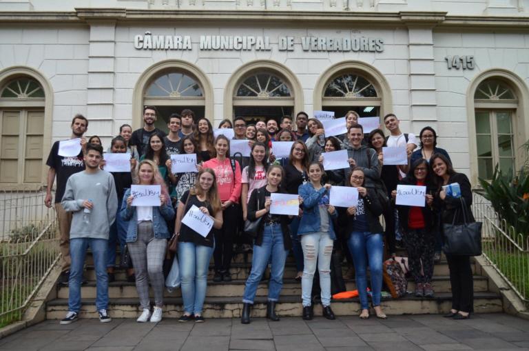 Diversos estudantes segurando cartazes em frente à Câmara de Vereadores de Santa Maria.