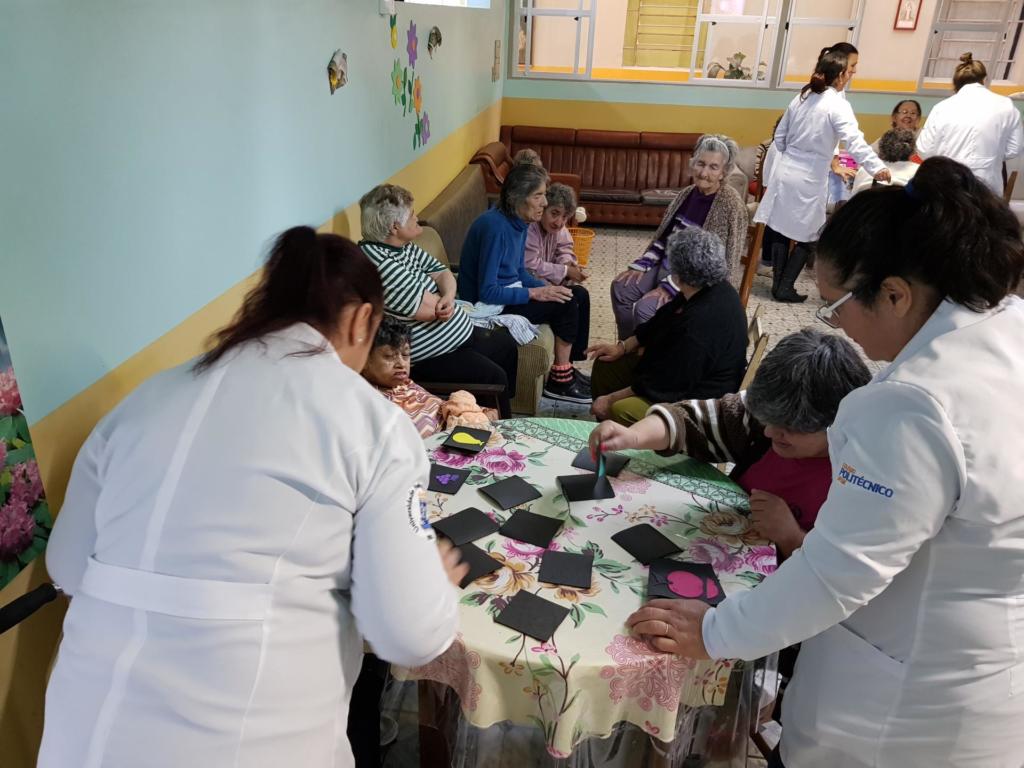 Alunas de jaleco de pé auxiliando idosos em atividades. Os idosos estão sentados em uma mesa redonda