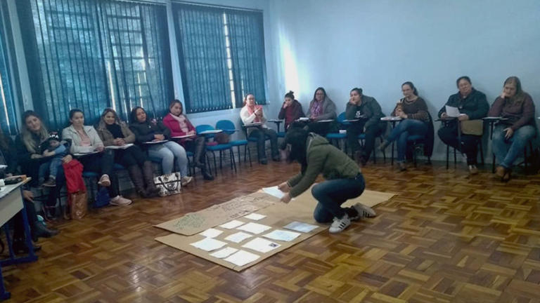 grupo de pessoas sentadas em círculo e ao centro outra pessoa colando folha de ofício em um cartaz.
